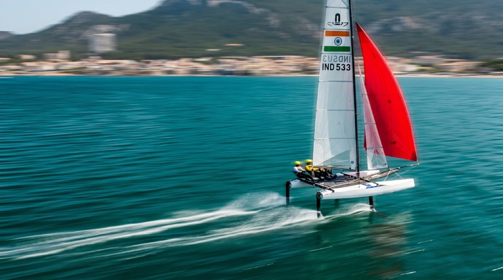 Foiling Nacra 17 with a red sail and Indian sail markings on turquoise water